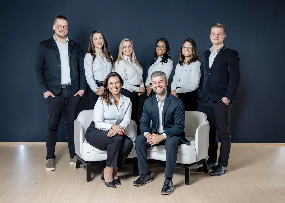 Equipe da Merco diante de uma parede escura: sete pessoas em pé ao fundo e os sócios Joice e Magnus sentados em poltronas à frente
