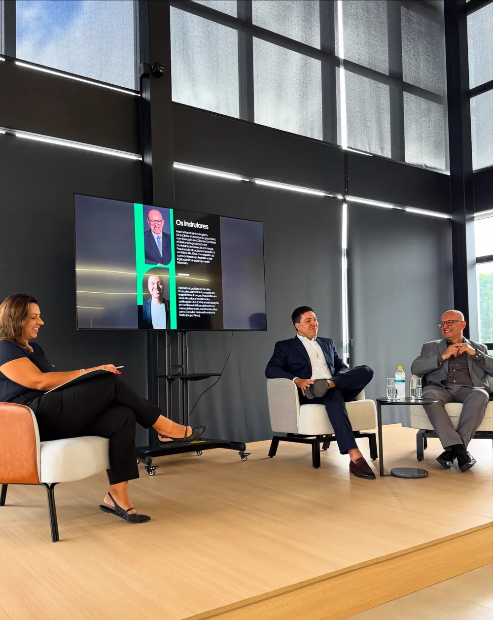 Painel da Merco em evento, com três palestrantes sentados no palco diante do público