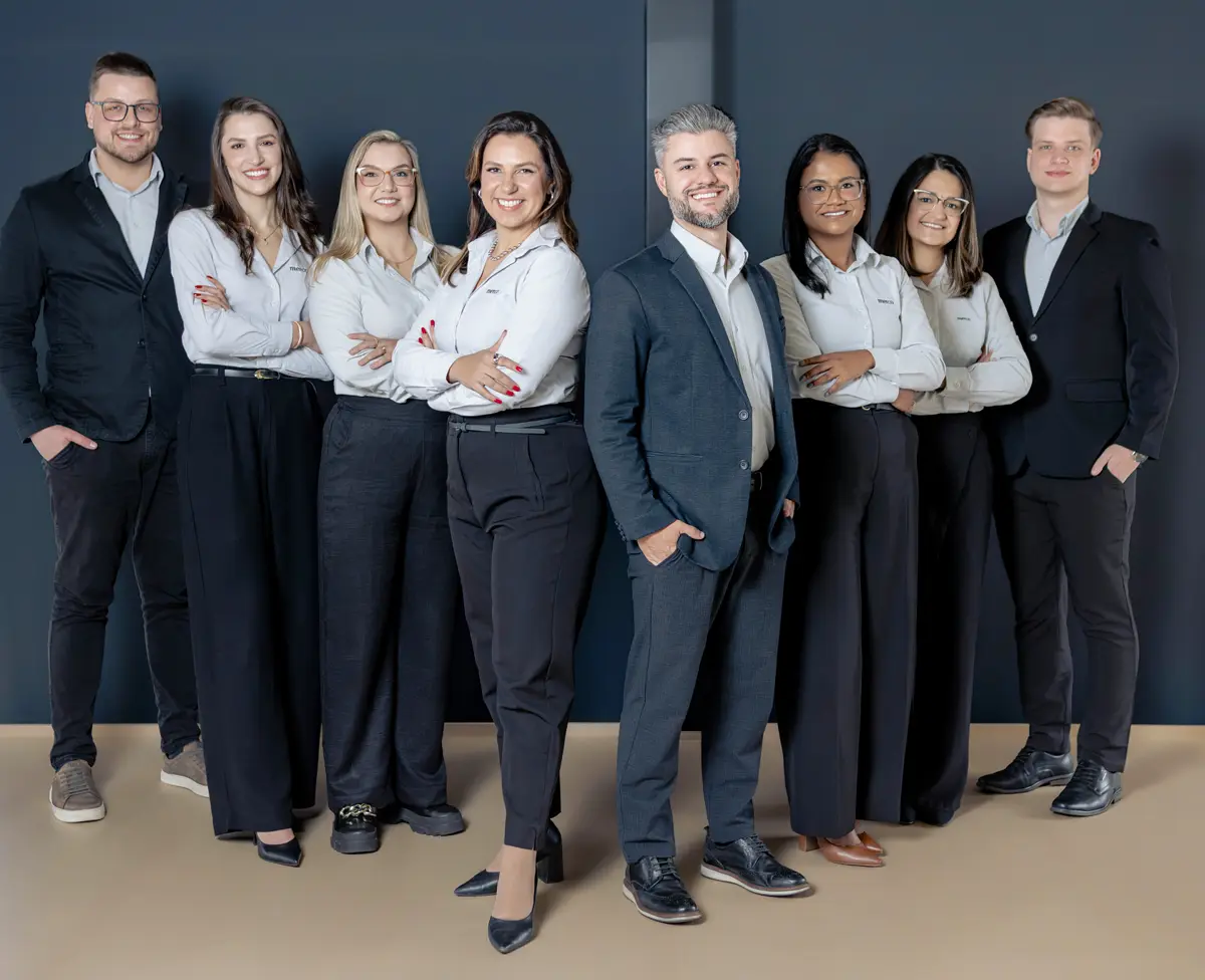 Equipe da Merco em pé, lado a lado, em frente a uma parede escura, com camisas claras e blazers
